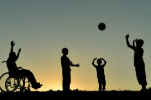 Silhouetten von Menschen beim Ballspielen im Abendlicht, eine Person im Rollstuhl